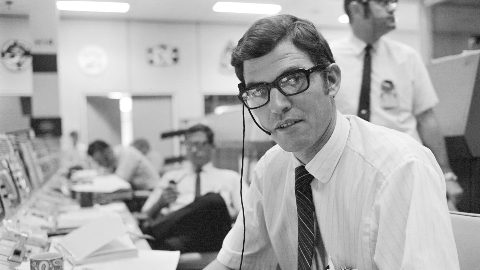 Black and white photo of a young flight controller in glasses and a headset, seated at a console in Mission Control with colleagues behind him