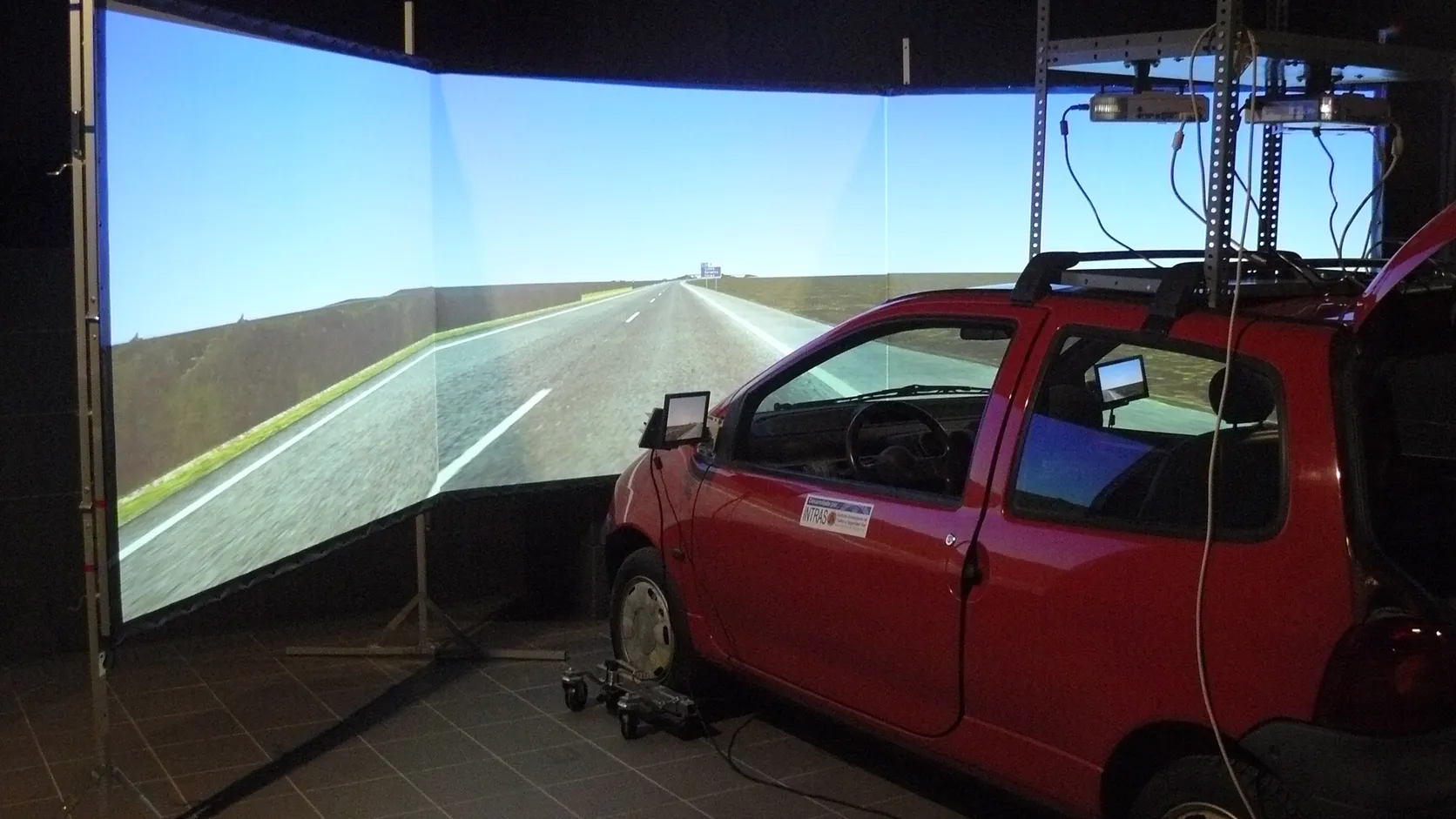 A red car parked in front of a wide three-panel projection screen showing a simulated highway